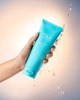 Hand holding a blue Tula skincare product with water droplets around it on a light background