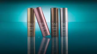 Four Tula Espresso hair eye balms arranged on a reflective teal surface, with three upright bottles and one tilted bottle in the center. The bottles are metallic gold, rose gold, and silver, 