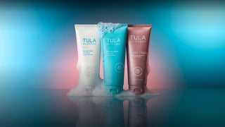 Three Tula Skincare tubes standing upright on a reflective surface with foam around their bases, set against a blue and pink gradient studio background.