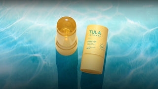 Tula Skincare Protect + Go Sunscreen Stick SPF 50, shown with the yellow stick container open and the matching yellow cap standing beside it on a blue water-textured background.