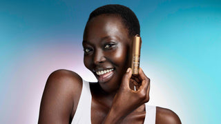 A smiling woman holds a TULA eye balm next to her face against a blue and purple gradient background.