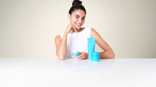 Woman with skincare products on a table against a plain background