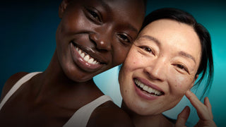 Two smiling women posing closely together against a teal-blue gradient background. The woman on the left has dark skin and is wearing a white strap top, while the woman on the right has light skin and is touching her cheek.