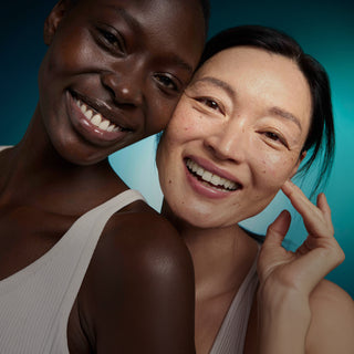 Two smiling women posing closely together against a teal-blue studio background. The woman on the left has dark skin and wears a white ribbed tank top, while the woman on the right has light skin and wears a pale pink top.
