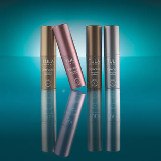 Four Tula Skincare eye drop bottles in metallic finishes (gold, rose gold, bronze, and silver) arranged on a reflective teal background.