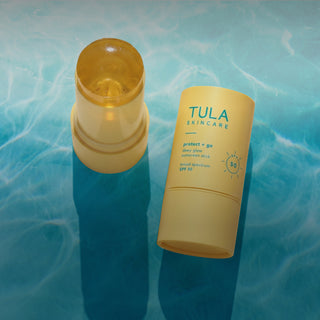 Tula Skincare Protect + Go Sunscreen Stick SPF 50, shown with the yellow stick container open and the matching yellow cap standing beside it on a blue water-textured background.