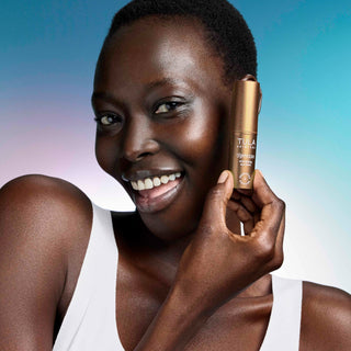 A smiling woman holds a TULA eye balm next to her face against a blue and purple gradient background.