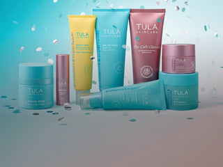 A collection of Tula Skincare products arranged together on a studio background with a teal-to-gray gradient and scattered confetti. The products include jars, tubes, and bottles in teal, pink, and yellow, with the brand name TULA SKINCARE visible on multiple items.