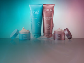 Four Tula Skincare products arranged on a reflective surface against a gradient studio background shifting from teal-blue on the left to pink on the right. The set includes two tall squeeze tubes and two open jars with cream visible.