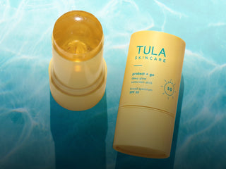 Tula Skincare Protect + Go Sunscreen Stick SPF 50, shown with the yellow stick container open and the matching yellow cap standing beside it on a blue water-textured background.