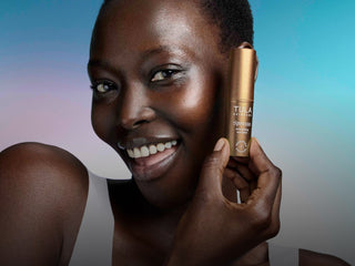 A smiling woman holds a TULA eye balm next to her face against a blue and purple gradient background.