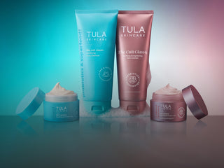 Four Tula Skincare products arranged on a reflective surface against a gradient studio background shifting from teal-blue on the left to pink on the right. The set includes two tall squeeze tubes and two open jars with cream visible.