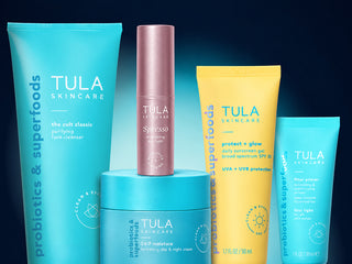 A set of five Tula Skincare products arranged together on a dark blue gradient background. The collection includes a large turquoise probiotics & superfoods tube, a smaller matching tube, a pink spresso skincare cream bottle, a yellow protect + glow sunscreen gel tube, and a turquoise eye cream tube, with one turquoise jar sitting on top of the larger tube.