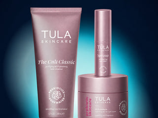 Three Tula Skincare products in matching metallic rose-pink packaging: a large tube of The Cult Classic facial cleanser, a smaller bottle of Spesso toner, and a jar of Prebiotic Skincare cream, arranged against a dark blue gradient background.