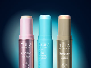 Three Tula Skincare eye balm tubes standing upright against a dark blue gradient background. The left tube is lavender with a pale pink cap and reads 'Spresso energizing eye balm.