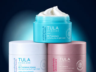 Three open jars of Tula Skincare creams stacked in a pyramid on a dark blue background. The top jar is turquoise with white cream visible inside, and the two bottom jars are white and dusty pink with their creams covered.