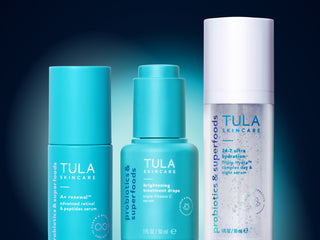 Three Tula Skincare skincare bottles in a row against a dark blue gradient background. The left bottle is a turquoise blue serum, the middle bottle is a matching turquoise blue treatment drop, and the right bottle is a white probiotic and superfoods spray with blue text.