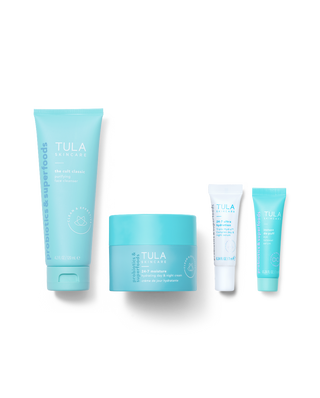 Four Tula Naturals skincare products in a row against a white background: a large light blue tube of probiotics & superfoods, a matching light blue jar of clay mask, a small white tube of face cleanser, and a light blue tube of eye cream.