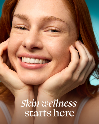 Close-up of a smiling woman with red hair resting her head on her hands, with the text 'Skin wellness starts here' overlaid at the bottom against a teal background.