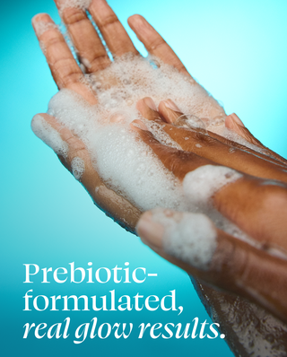 Close-up of two hands covered in white foam against a bright aqua background, with the text 'Prebiotic-formulated, real glow results.' overlaid at the bottom.